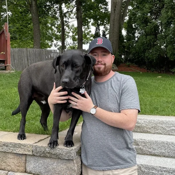 man with baseball cap standing next to black dog